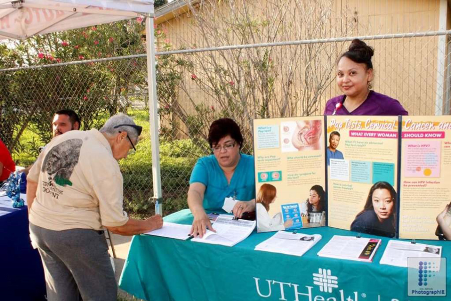 Photograph of two women dressed in scrubs standing at a stall promoting health screening. A member of the public is leaning over the table writing in a sign-up sheet.