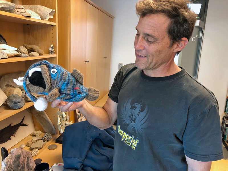In his UC Santa Barbara office, Kevin Lafferty holds a plush-toy anglerfish knitted by former post-doctoral researcher Julia Buck. The stuffed animal is sufficiently anatomically correct to show how the tiny, parasitic male anglerfish, colored red, implants himself into the female’s body. The male feeds off his mate’s circulatory system while supplying sperm. The shelves are filled with marine fossils. 