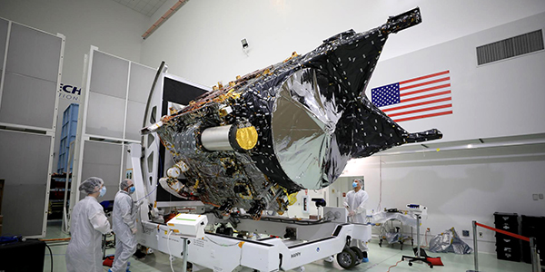 Workers dressed in white clean suits stand around the chassis of the Psyche spacecraft in a facility adorned with a large American flag.