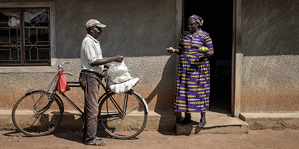A participant in an experiment on giving people a long-term universal basic income stands outside her house holding oranges she has bought from a man leaning on bicycle in Kenya. 