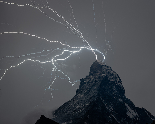 Photo of bright, jagged lightning bolts striking a black mountain peak against a night sky.