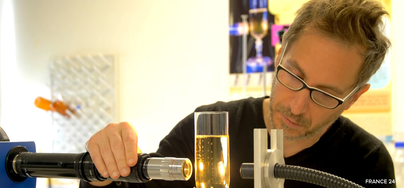 Photograph of a man with glasses and light beard and moustache wearing a black sweater; he is leaning over a high-speed camera that is trained on a glass of champagne so that he can study the bubbles that are forming.
