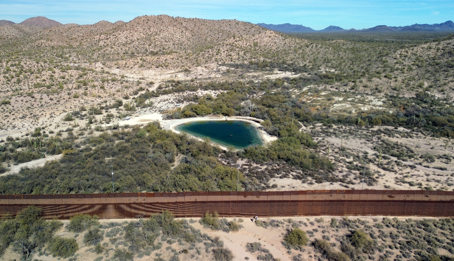 Aerial image showing an arid terrain, with green bushes and, in the middle, a body of water known as the Quitobaquito spring. The terrain is traversed from side to side by a wall, preventing access to the spring from the southern side.