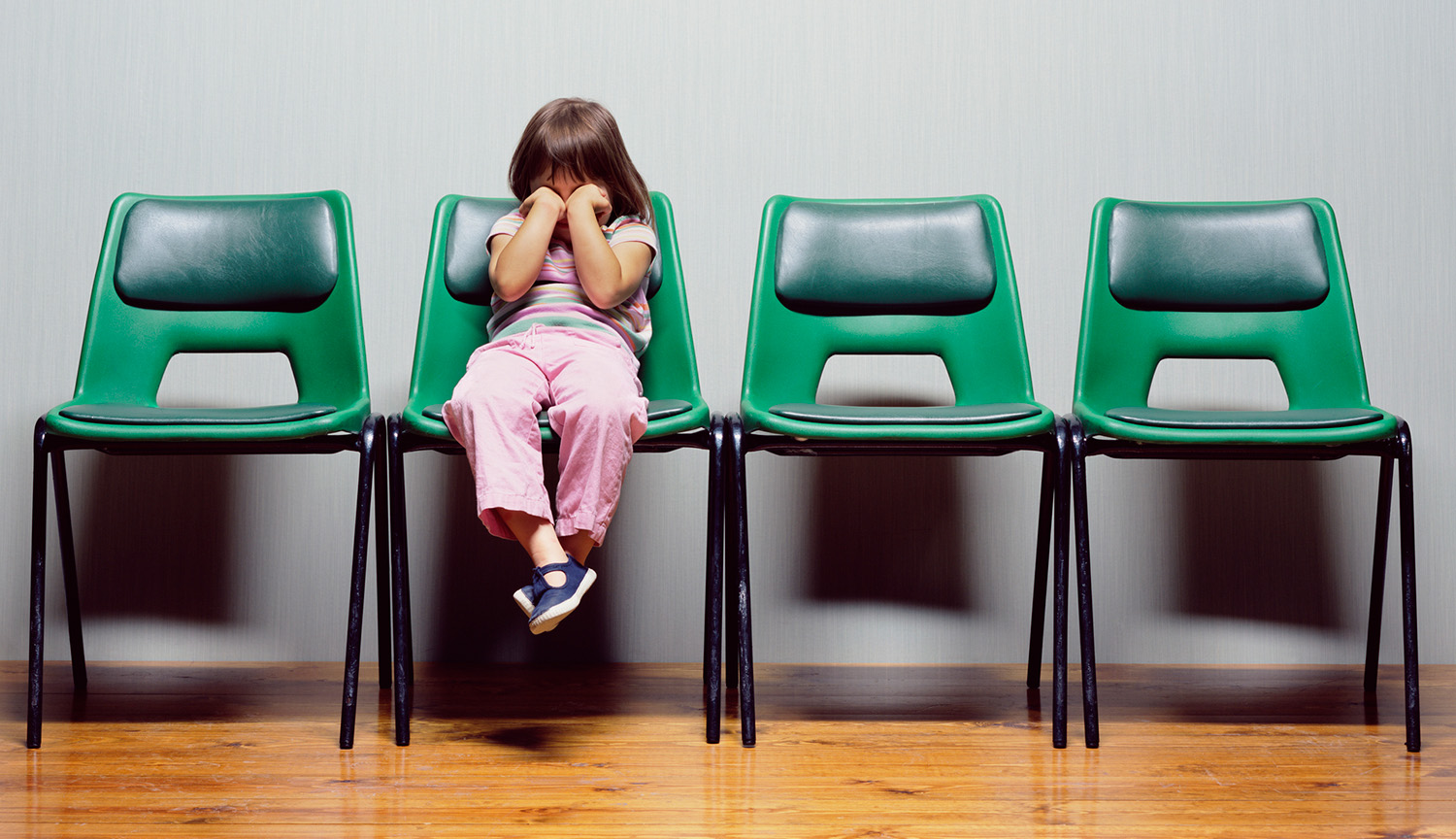 Photograph of a small child sitting in a waiting-room chair with fists covering their face.