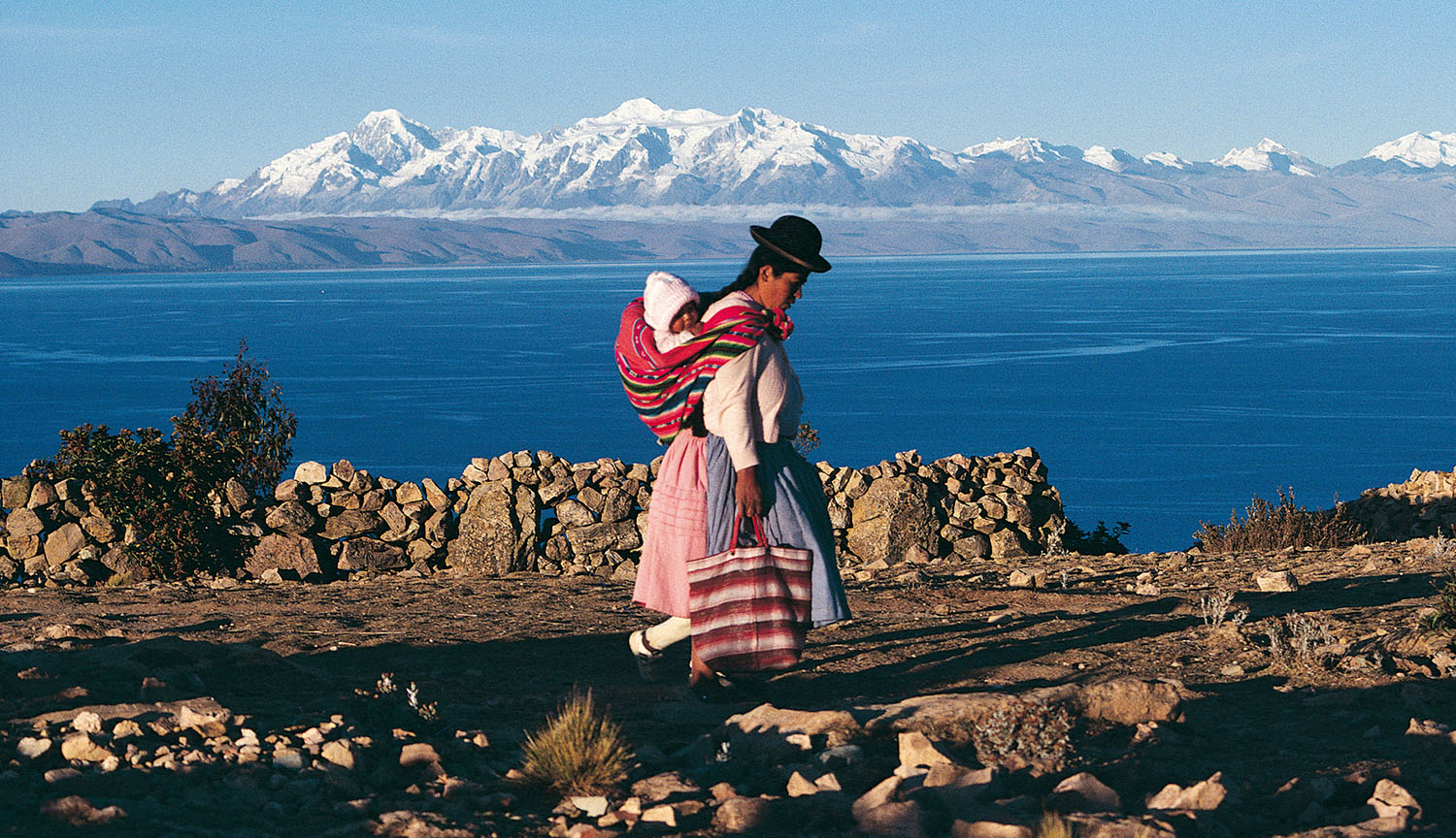 An indigenous woman in traditional dress with a baby on her back stands in front of a lake, with high mountains in background.