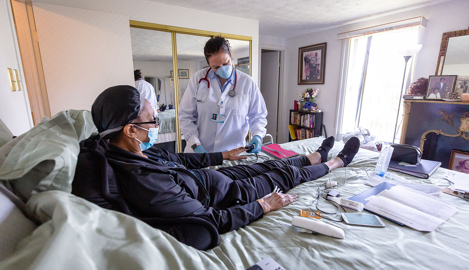 Photograph of a woman lying on her bed in her home while a nurse in white coat and mask checks her vital signs.