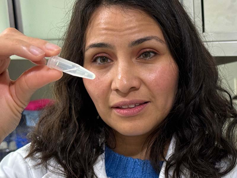 A woman with black hair, dressed in a lab coat, holds a pipette.