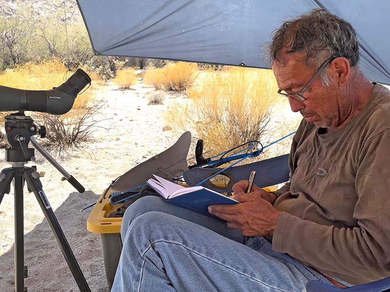 A man sitting in the shade of a tent takes notes in a notebook.