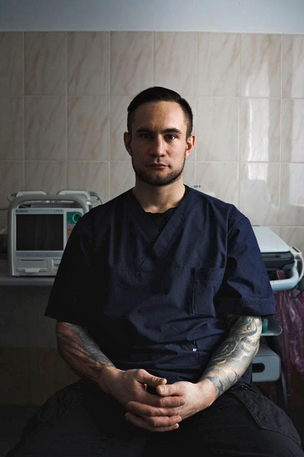 Ukrainian surgeon A serious young man in dark blue scrubs, his tattooed arms folded in front of him, in a hospital room.
