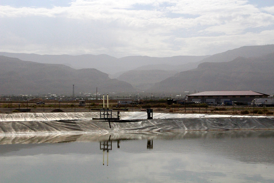 A large reservoir of dirty-looking water is backed by white slanted walls. There are mountains in the distance and clouds in the sky. 