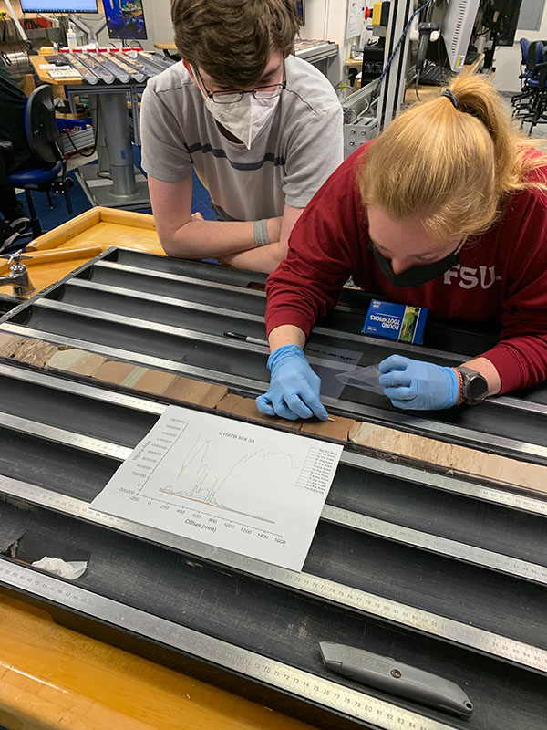 Sampling sediment cores A woman leans over a long core of sediment in metal housing; you can see different layers of sediment in different colors. She is wearing gloves and sampling the sediment while a second individual looks on.