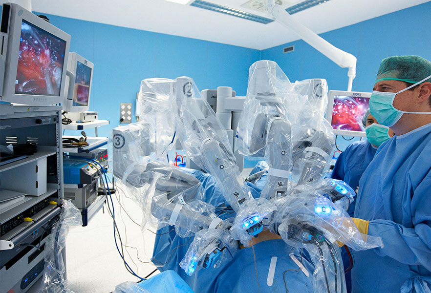 The robo-surgeon is in Several plastic-draped robotic arms tend to a patient while the surgeon gazes at a nearby screen.