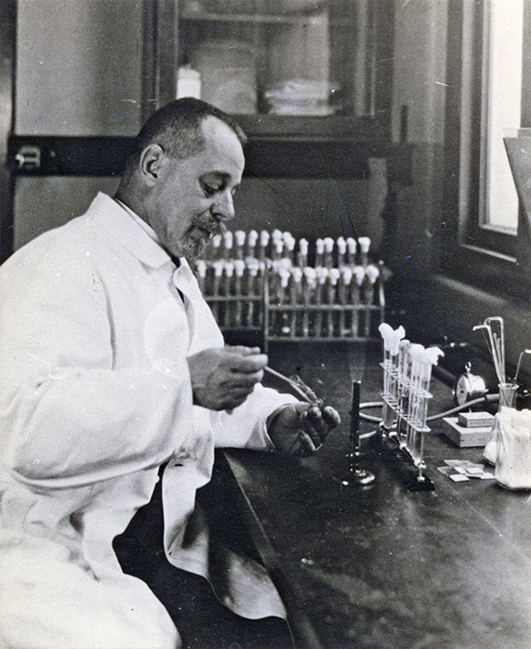 Vintage photo in which a man in a white coat sits at a laboratory bench with racks of test tubes.