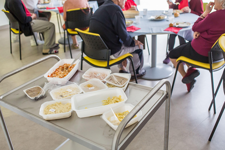 Photo of food in cartons on a tray; nearby are tables with nursing home residents sitting around them. The foods are bland shades of brown and beige.