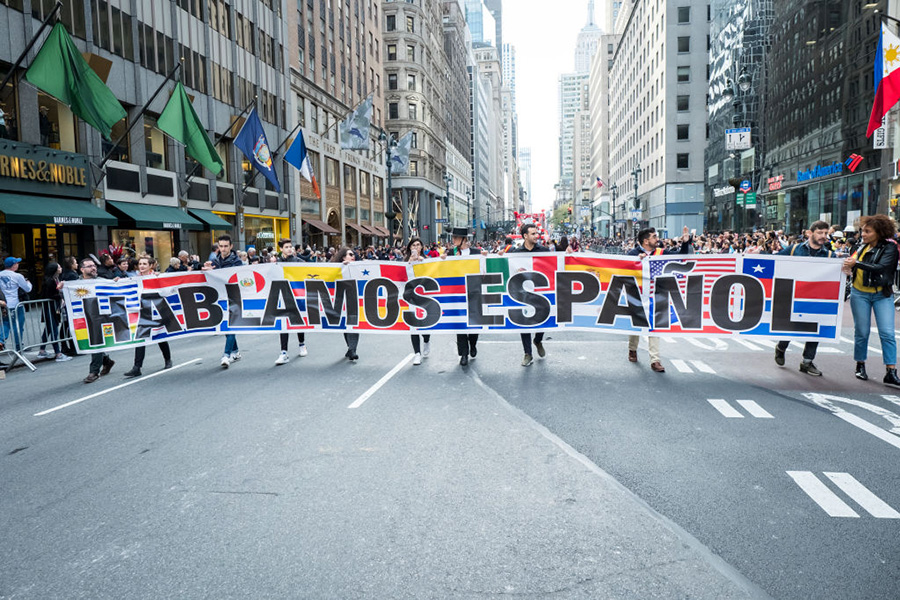 People march in the middle of an avenue carrying a sign with flags of Hispanic countries and written over them the phrase “Hablamos español” (we speak Spanish).