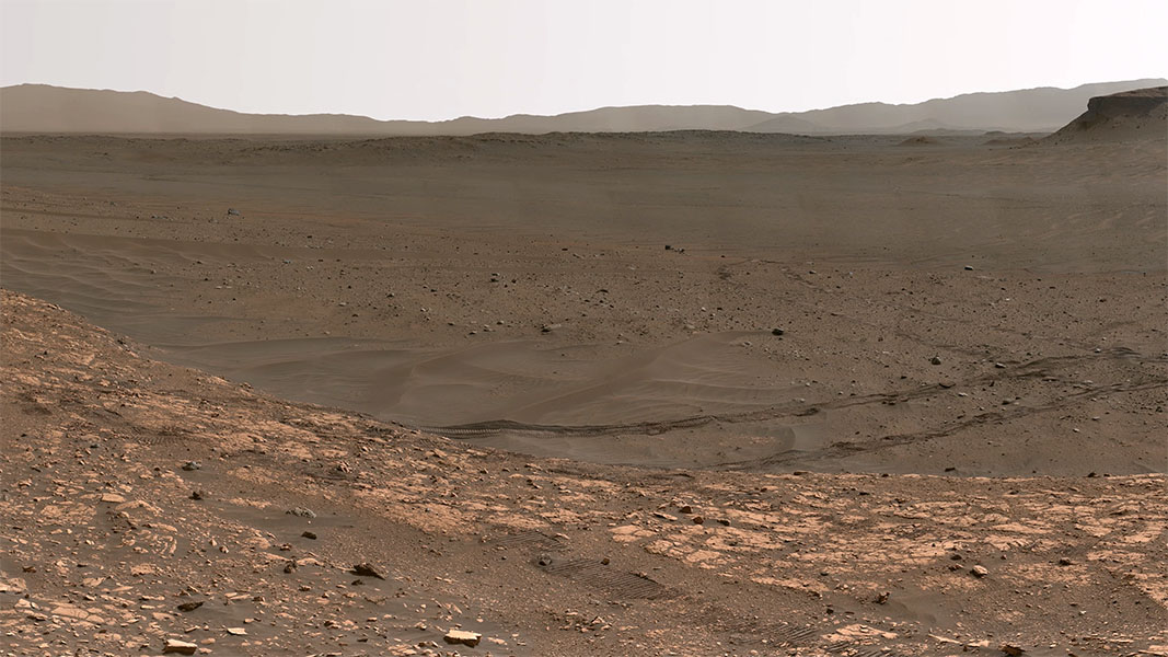 A view from the Mars rover shows a dry reddish valley with rocks in the foreground and mountains in the far distance.