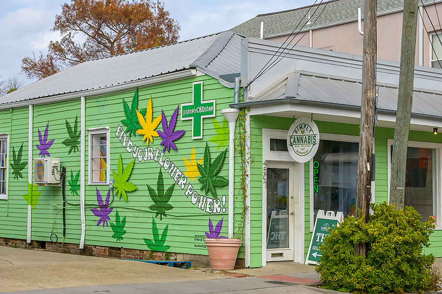 Photo shows a brightly painted small building with marijuana leaves in yellow, purple and green decorating the side of the building. A green cross symbol and a front sign adorn the store.