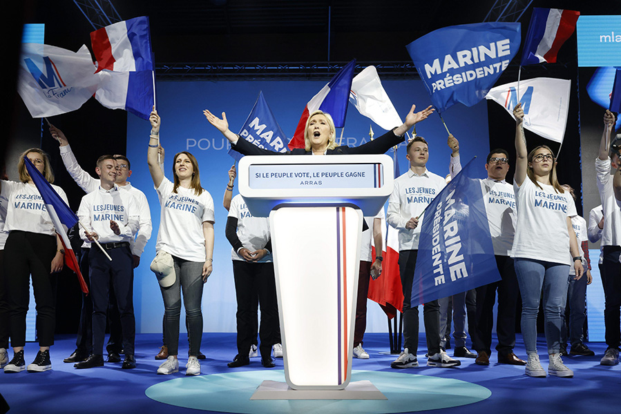 A blond woman stands in front of a lectern with her arms cast wide in the air while people with “Marine” T-shirts wave French flags and ones calling for her to be president. Written on the lectern is “si le peuple vote, le peuple gagne” — “if the people vote, the people win.”