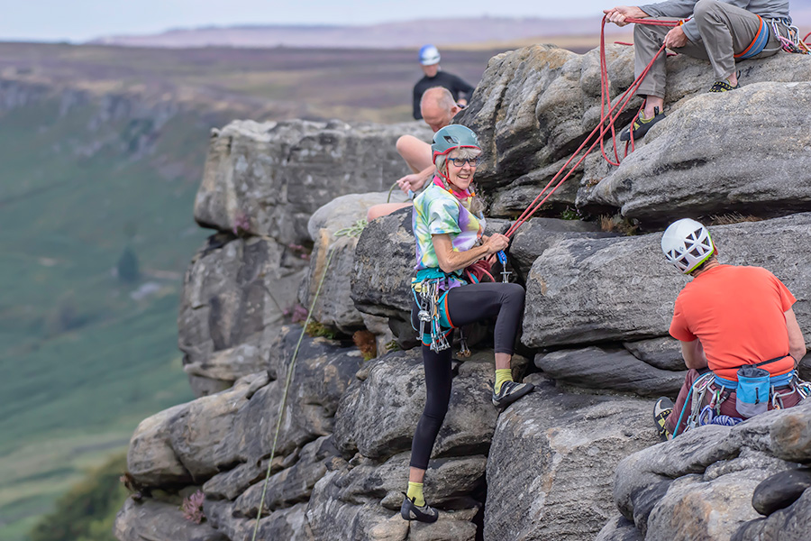 A mature woman stands at the top of a cliff having completed a rock climb. She is smiling.