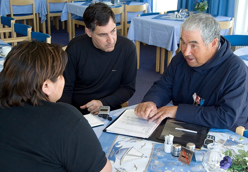 Three people are seated at a table. One of them, an older man, is talking and referring to a notebook, while the other two listen intently.