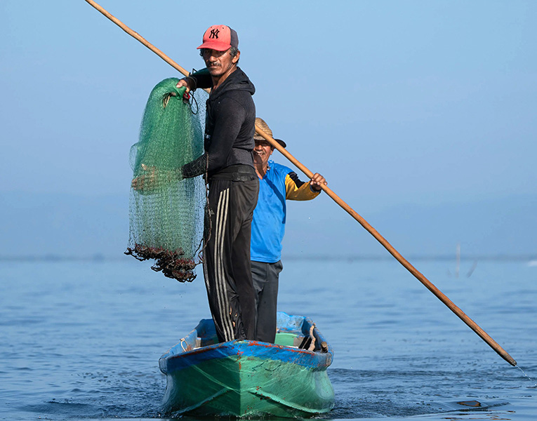 Two men travel in a small boat. The man in the background rows, while the man in front holds a net with freshly caught shrimp.