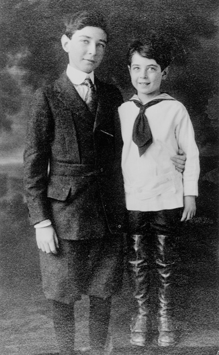 Una antigua foto de estudio en blanco y negro muestra a dos chicos, uno más joven con traje de marinero y botas largas, el mayor más alto con traje de lana.