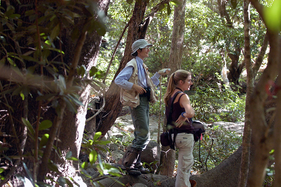 Photo of two women standing among trees and vegetation looking off into the distance. One of them, Susan Perry, is wearing a khaki hat and holding a logbook.