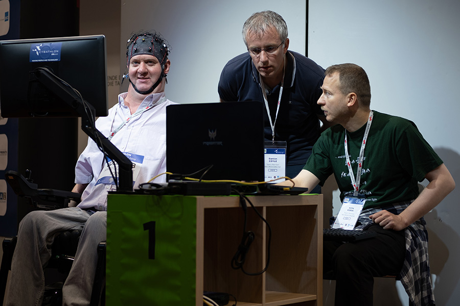 A man with an EEG headset looks at a computer screen, two other men look on and at a second screen.