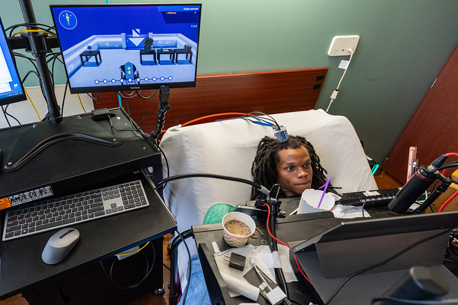 A man in a bed with an implant coming out of the top of his head uses his thoughts to control an avatar on a computer screen.