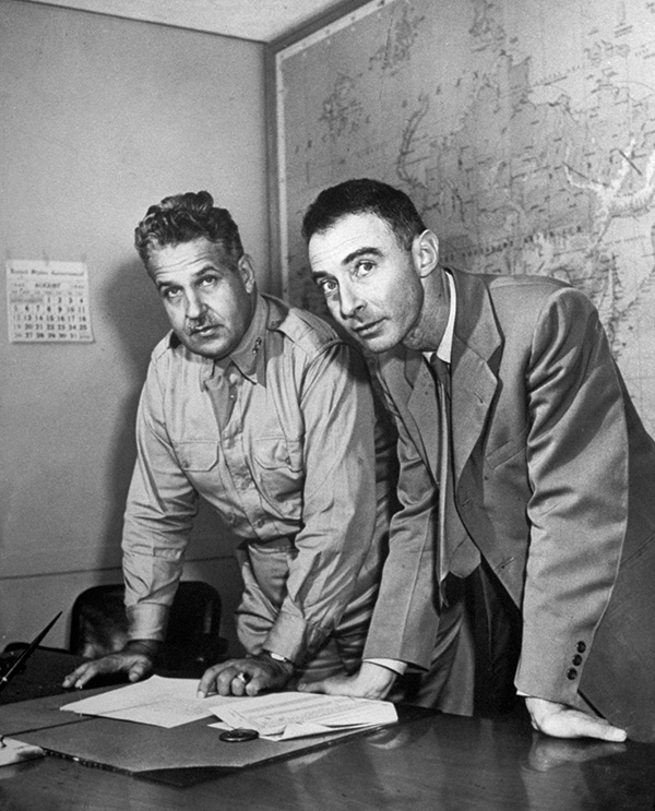 Una foto en blanco y negro de dos hombres, uno con uniforme militar y el otro con traje, de pie ante un escritorio, con un mapa detrás de ellos.