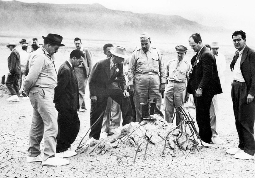 Una foto en blanco y negro muestra a un pequeño grupo de hombres con trajes y uniformes rodeando un montículo de tierra con barras de acero dobladas asomando por él, sobre un fondo desértico.
