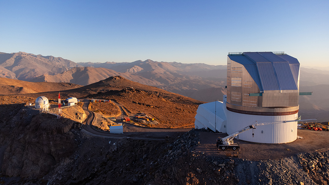 A sunset view of Rubin Observatory atop its Chilean desert mountain site; mountain ridges recede into the distance under a blue sky. 