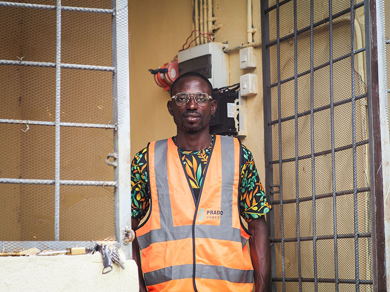 Photograph of a Nigerian man wearing an orange safety vest over a brightly patterned shirt. He is standing in a doorway.