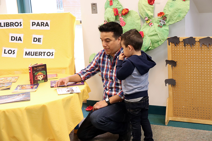 A boy stands next to a man that is kneeling in front of a table with children’s books. In the background there is a sign that says in Spanish “Libros para día de los muertos” (Books for the Day of the Dead).