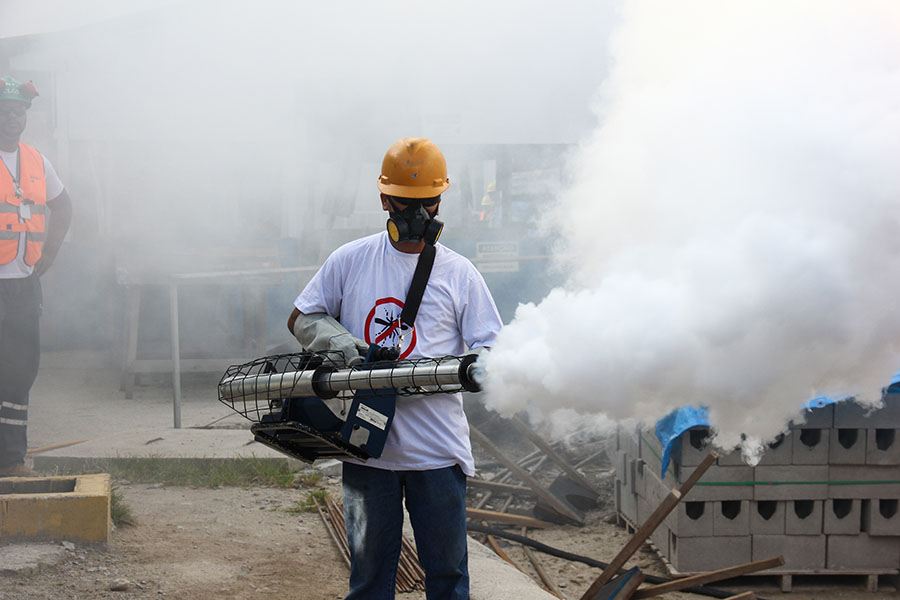 Photo of a man in protective gear fumigating an area; there are billows of smoke coming out of the fumigator.