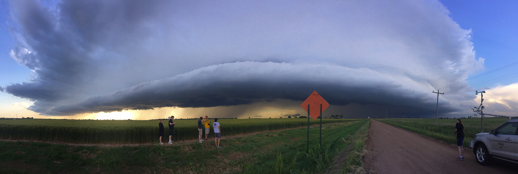 Cinco estudiantes universitarios de pie en el borde de un maizal observando una gigantesca nube de tormenta baja. 