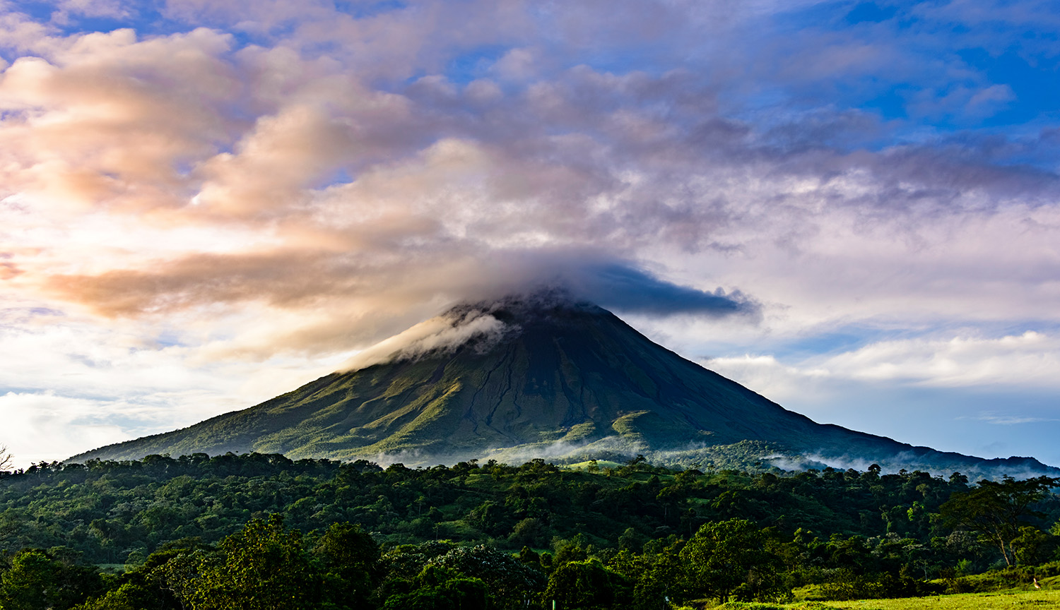 An impressive cone-shaped volcano, with clouds over its summit, a lake on the side and green vegetation in front.